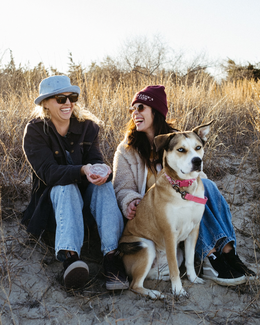 Two people sitting on a sandy beach with a dog, wearing hats and sunglasses.